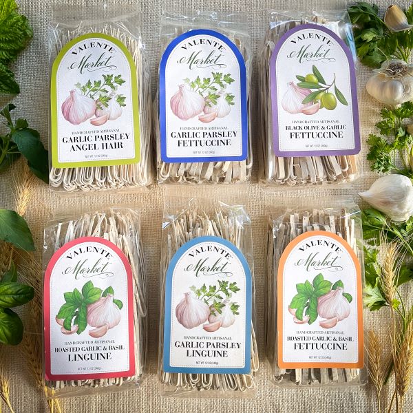 Six 12 oz packages of pasta with colorful labels. One Garlic Parsley Linguine, one Roasted Garlic & Basil Linguine, one Black Olive & Garlic Fettuccine, one Roasted Garlic & Basil Fettuccine, and one Garlic Parsley Fettuccine, one Garlic Parsley Angel Hair.