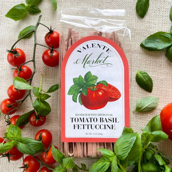 Tomato basil fettuccine with a bright red border label in a clear bag, surrounded by fresh tomatoes and basil
